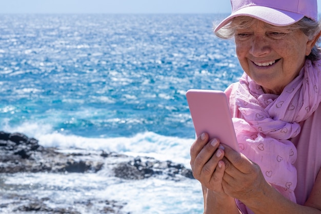 Lachende senior vrouw, roze gekleed, met behulp van telefoon op zee in winderige dag. senioren genieten van vakantie en schoonheid in de natuur