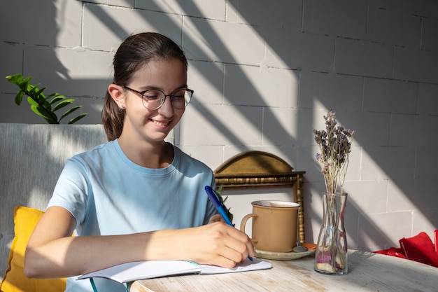 Lachende studente schrijft huiswerk met een pen in een notitieboekje in een café. Portret van een blank brunette meisje met een bril en een blauwe blouse in een café met diagonale schaduwen. Terug naar schoolconcept.