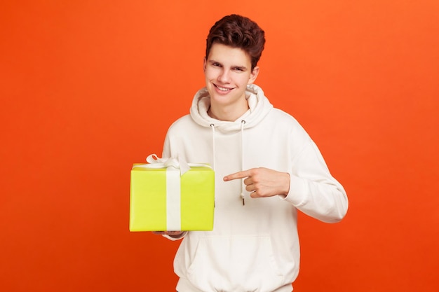 Lachende tiener in casual stijl sweatshirt wijzende vinger op grote geschenkdoos in de hand promoties en kortingen in uw tarief mobiele communicatie Indoor studio opname geïsoleerd op oranje achtergrond