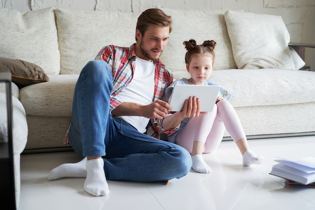 Lachende vader en dochter zittend op de vloer in de woonkamer met digitale tablet, lesgeven.