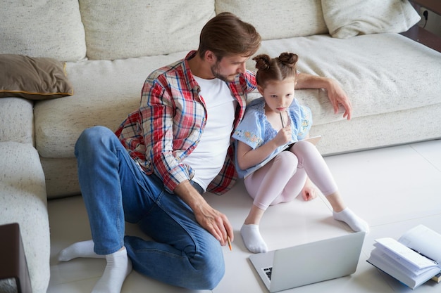 Lachende vader en dochter zittend op de vloer in de woonkamer met laptop, lesgeven.