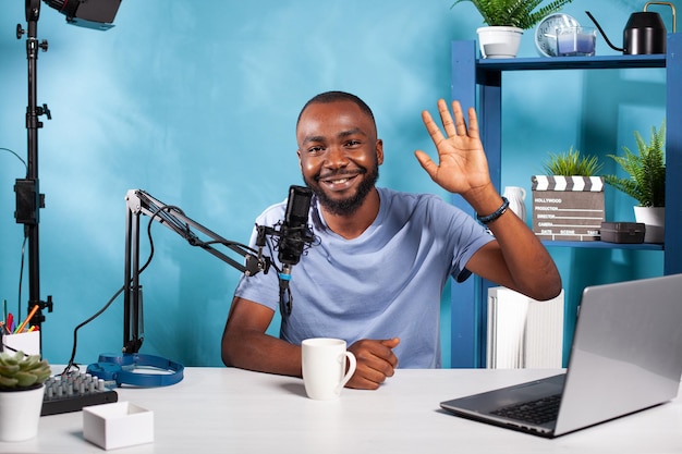 Lachende vlogger zwaaiend hallo zittend aan bureau in opnamestudio met professionele microfoon en videolicht. Contentmaker doet handgebaar voor de installatie van audiopodcasts.