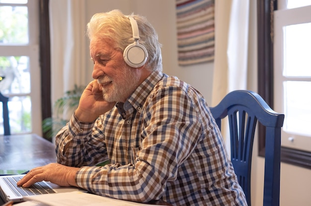 Lachende volwassen bebaarde man met koptelefoon aan tafel zitten met laptop na online cursus