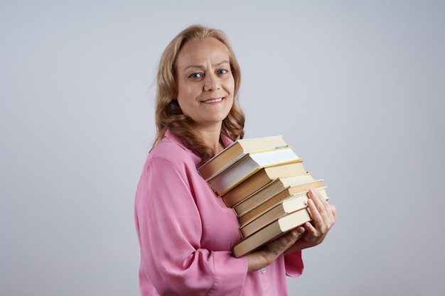 Lachende volwassen vrouw leraar, bibliothecaris in roze shirt, met een stapel boeken, op zoek. Leerconcept.