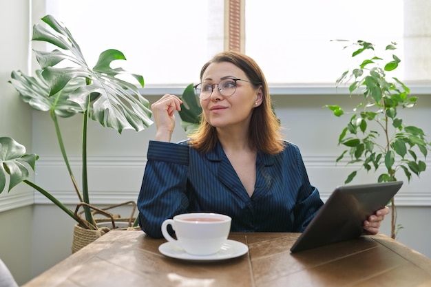 Lachende volwassen vrouw ontbijten thuis in pyjama met kopje thee zittend aan tafel lezen van digitale tablet. Gelukkig wijfje van gelezen post, nieuws. Positieve emoties, informatie, het leven, mensen uit de jaren 40