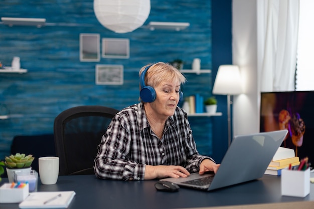 Lachende volwassen vrouw ontspannen met draagbare computer Gepensioneerde senior vrouw hoofdtelefoon dragen tijdens het gebruik van laptopcomputer in huis woonkamer.