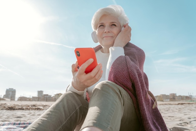 Lachende vrolijke oudere dame in draadloze koptelefoon en een smartphone in de hand zittend op het zand