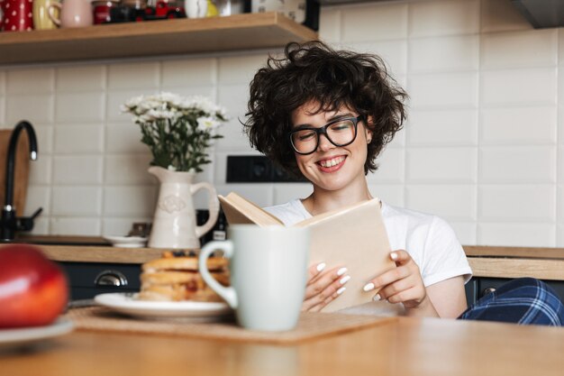 Lachende vrolijke vrouw lekker ontbijten zittend in de keuken thuis, een boek lezen