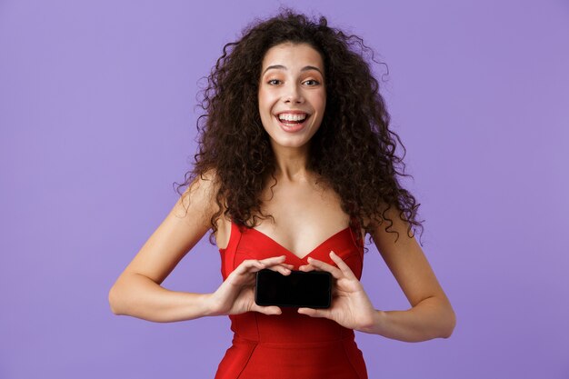 lachende vrouw, gekleed in rode jurk met zwarte smartphone, geïsoleerd over violette muur