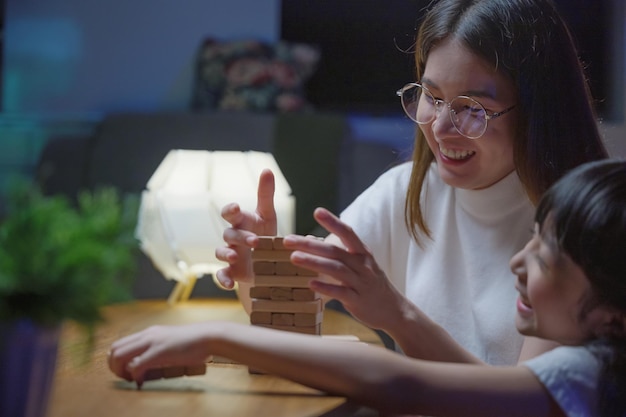 Lachende vrouw helpt kinderen spelen te leren bouwen constructeur van houten blokken Aziatische jonge moeder speelspel in houtblokken met haar dochtertje in huis woonkamer 's nachts voordat ze naar bed gaat