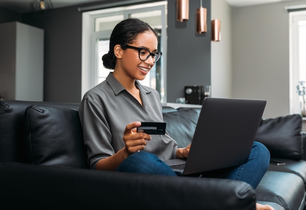 Lachende vrouw in bril met een creditcard die naar een laptop kijkt terwijl ze op een bank in de woonkamer zit