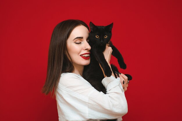 Lachende vrouw in lichte vrijetijdskleding staat op een rode achtergrond met een zwarte kat in haar handen