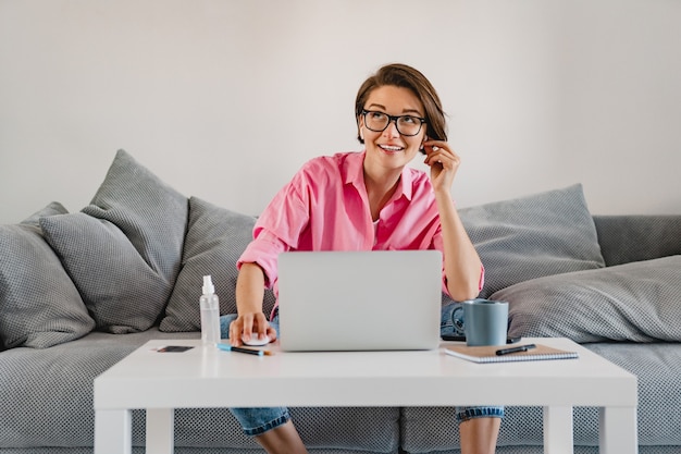 lachende vrouw in roze shirt ontspannen zittend op de bank thuis aan tafel online werken op laptop vanuit huis