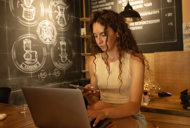 Lachende vrouw maakt gebruik van laptopcomputer, zit bij coffeeshop met kopje cappuccino en dessert