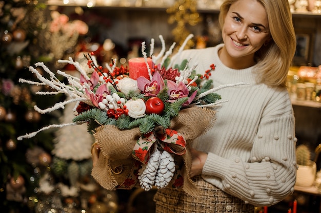 Lachende vrouw met een Kerstmissamenstelling met roze orchideeën, witte rozen, dennentakken, rode appel en kaars in de zak in de bloemenwinkel