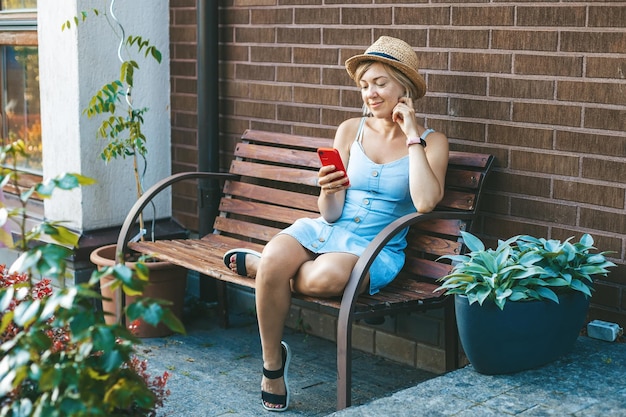 Foto lachende vrouw met strohoed die smartphone gebruikt terwijl ze op een bankje in de tuin zit