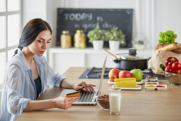 Lachende vrouw online winkelen met behulp van computer en creditcard in de keuken