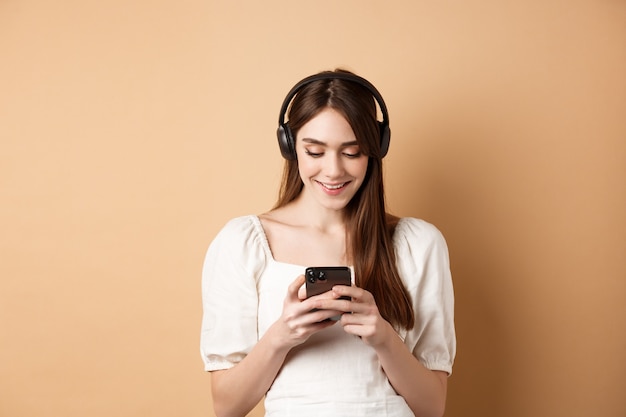 Lachende vrouw SMS-bericht op telefoon en luisteren muziek in draadloze hoofdtelefoons, kijken naar video op smartphone, beige achtergrond.