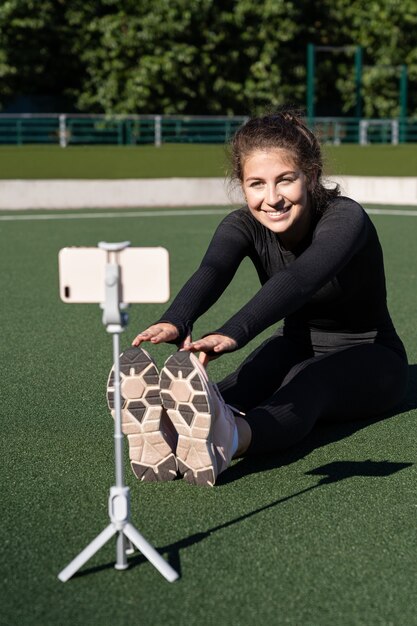 Lachende vrouw vlogger in sportkleding zittend op kunstgras buiten