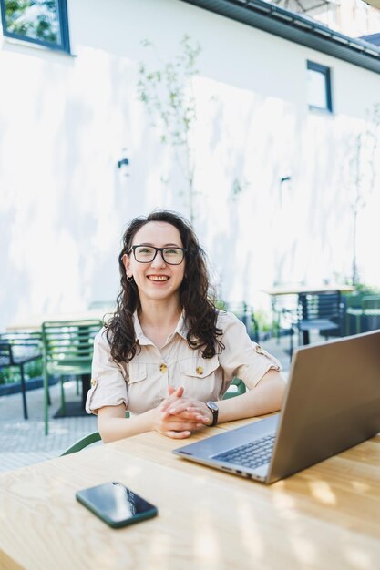 Lachende vrouw zittend aan terrastafel met laptop praten over telefoon en kopje koffie Lachende vrouw in glazen genieten van telewerken in café of online studeren