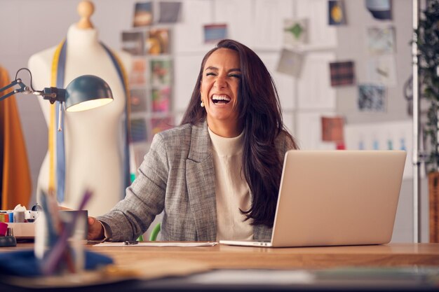 Lachende vrouwelijke modeontwerper in studio bezig met schetsen of documenten aan bureau met laptop
