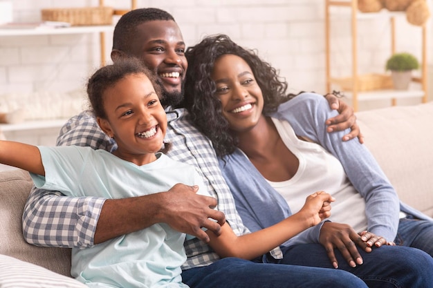 Foto lachende zwarte moeder, vader en dochter ontspannen op de bank thuis, samen genieten van tijd.