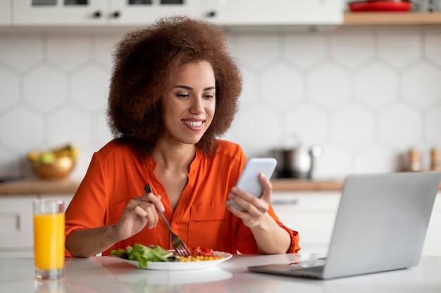 Lachende zwarte vrouw die smartphone en laptop gebruikt tijdens de lunch in de keuken