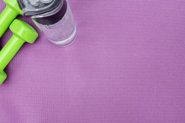 Ladie's dumbbells and water bottle over purple fitness mat top view Sport concept