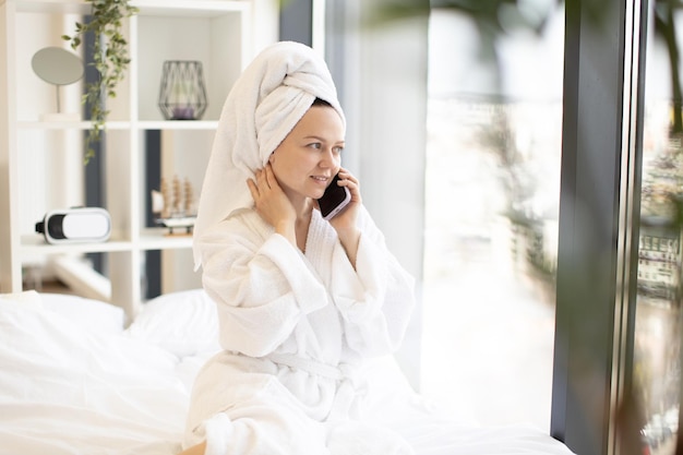 Lady in bathrobe talking on smartphone in bed at home