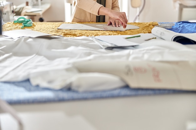 Lady dressmaker draws pattern on yellow fabric sheet with chalk and detail at large cutting table 