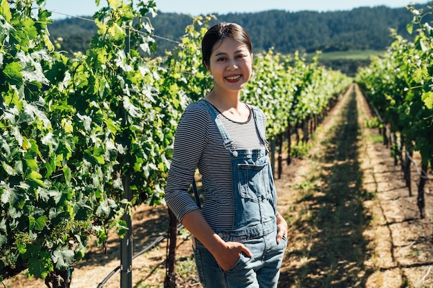 夏の晴れた日にブドウ園でブドウを収穫する農家の女性。ぶどうワイナリーのアジアの若い女の子のワインメーカーは、カメラに自信を持って微笑んでいます。うれしそうに農場に立っている美しい女性。