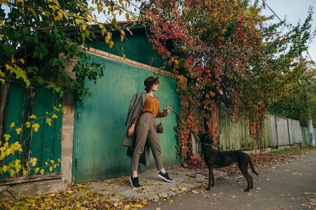 Signora in abiti alla moda si trova sulla strada contro una casa di campagna e un paesaggio autunnale