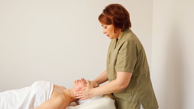 La signora in cappotto verde massaggia la schiena e il collo del visitatore del salone di bellezza dopo le operazioni cosmetiche sul divano bianco nel reparto sotto una luce intensa