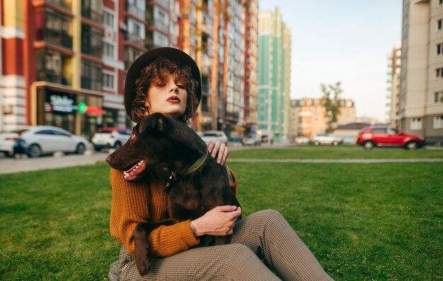 Lady in hat sits in yard on lawn with dog in her hands and poses at camera on cityscape