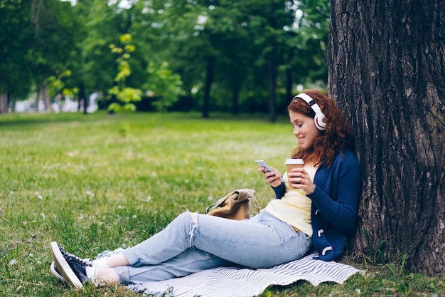 公園でコーヒーを飲みながらスマートフォンを使用して音楽を楽しんでいるヘッドフォンの女性