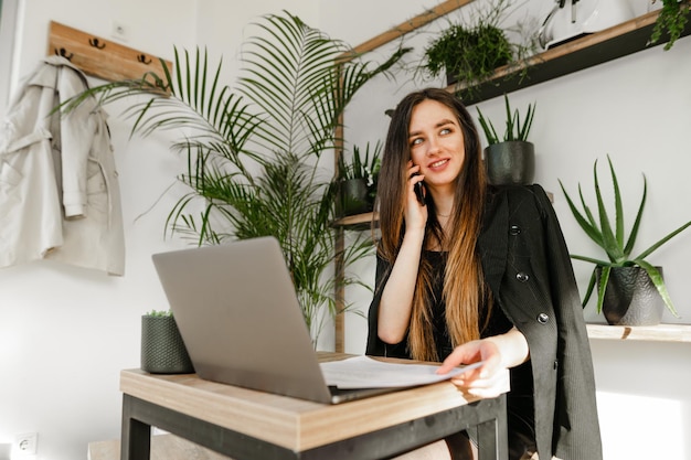 lady in a jacket sits at table in a cozy cafe with laptop communicates on the phone and looks away