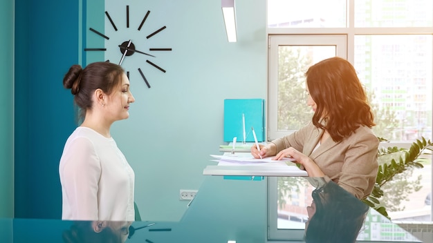 Офис-менеджер леди в белой блузке показывает регистрационную книгу клиентке, стоящей на стойке регистрации в современной больнице, вид сбоку