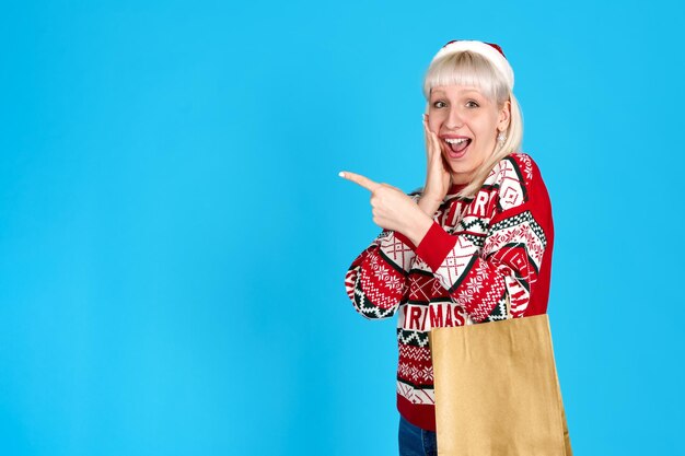 Lady in red Christmas outfit holding a shopping bag pointing to copyspace on blue studio backdrop