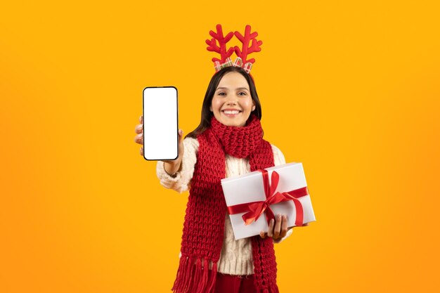 Lady showing phone screen holding xmas gift box yellow background