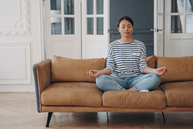 Lady sitting on sofa in lotus asana and meditating in living room Stress relief and relaxation