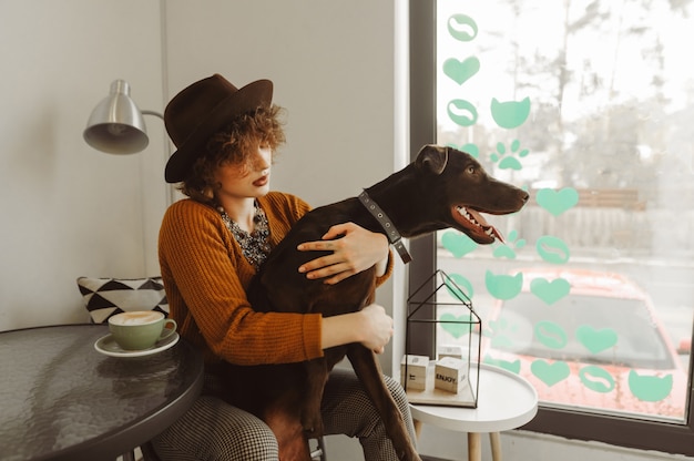 Lady in stylish clothes playing with a dog in a cozy coffee shop by the window