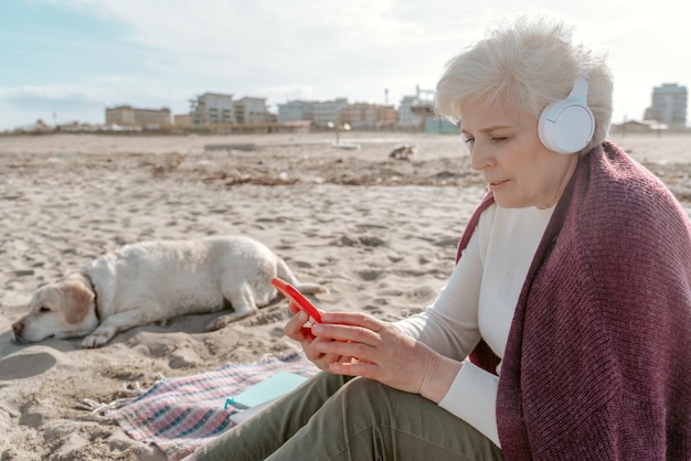 ワイヤレス ヘッドフォンを装着した女性が犬のそばの砂の上に座り、スマートフォンの画面を見つめている