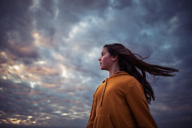 Foto lage hoek van een meisje met lang haar dat wegkijkt terwijl het tegen een bewolkte hemel staat