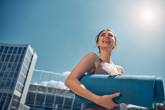 Lage hoek van een vrolijke jonge vrouw die met beide handen een opgerolde yogamat vasthoudt