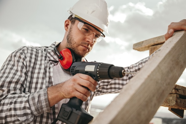 Lage hoek van ernstige bebaarde mannelijke werknemer in geruit overhemd en beschermende bril en veiligheidshelm boren houten structuur, met professioneel instrument tijdens het werken op de bouwplaats