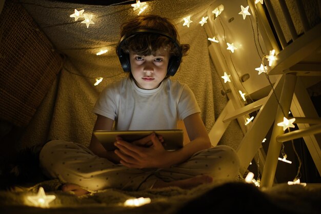 Lage hoek vol lichaam van jongen in pyjama en draadloze koptelefoon kijken naar video op tablet zitten in het spelen tent versierd met gloeiende krans