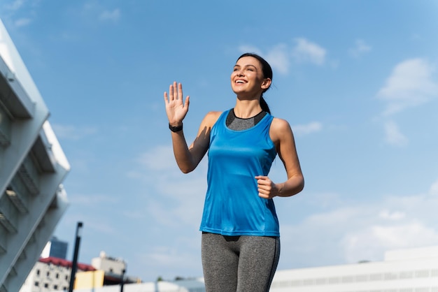 Lage hoekmening van de gelukkige gemotiveerde vrouw die met haar hand naar iemand zwaait en vrolijk glimlacht tijdens het joggen Blauwe hemelachtergrond