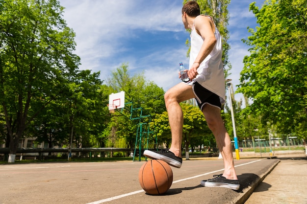 Lage hoekmening van jonge atletische man staande op zijlijnen van basketbalveld met voet bovenop bal voorkomen dat het wegrolt, en kijken naar bord en mand