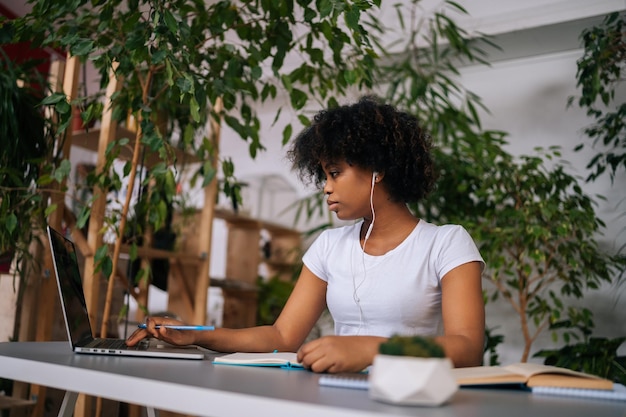Lage hoekmening van zwarte vrouwelijke student die oortelefoons draagt en notities schrijft die kijken naar webinar die online studeert met een laptop die aan tafel zit
