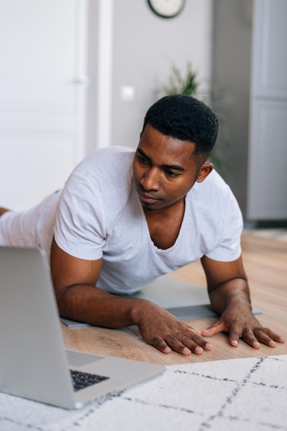 Lage hoekopname van een jonge Afro-Amerikaanse man die training in horizontale plankpositie uitoefent met behulp van een laptop om yoga te kijken en een digitale video-tutorial te trainen.